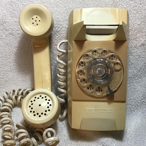 May include: A cream-colored wall-mounted rotary telephone with a matching handset and a coiled cord. The device features a circular dial with numbers and letters, and the text GTE AUTOMATIC ELECTRIC is printed on the lower front section.