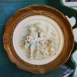 May include: A decorative, antique wall plate with a gold-colored, ornate frame. The center features a bas-relief scene of two figures in period clothing, with a musical instrument and a scroll. The plate is off-white with pastel accents.