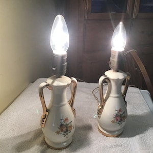 May include: Pair of white ceramic table lamps with gold accents and floral designs. The lamps have a classic, vintage style.