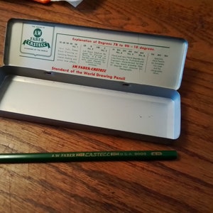 May include: A green and gold A.W. Faber-Castell pencil in a metal tin case. The tin has a label that reads "Standard of the World Drawing Pencil" and explains the different degrees of hardness for drawing pencils.