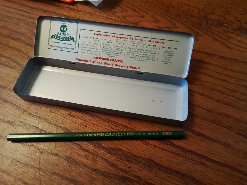 May include: A green and gold A.W. Faber-Castell pencil in a metal tin case. The tin has a label that reads "Standard of the World Drawing Pencil" and explains the different degrees of hardness for drawing pencils.