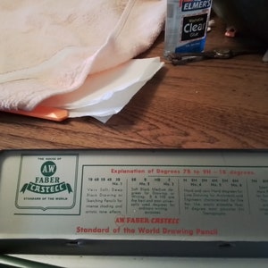 May include: A vintage metal tin box with a green and white label that reads "A.W. Faber-Castell Standard of the World Drawing Pencil". The label explains the different degrees of hardness for drawing pencils, from 7B to 9H.