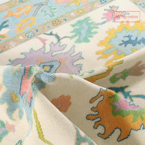 May include: Close-up of a cream-coloured rug with a detailed floral pattern. The rug features a border with light blue, orange, and green accents. The central design includes pastel-coloured floral and abstract shapes.