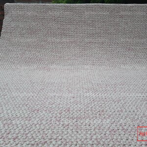 May include: A close-up of a white and pink textured rug. The rug is made of small, round balls of yarn. The rug is folded in half and the bottom edge is visible. The text "Red Rugs creations" is visible in the lower right corner of the image.