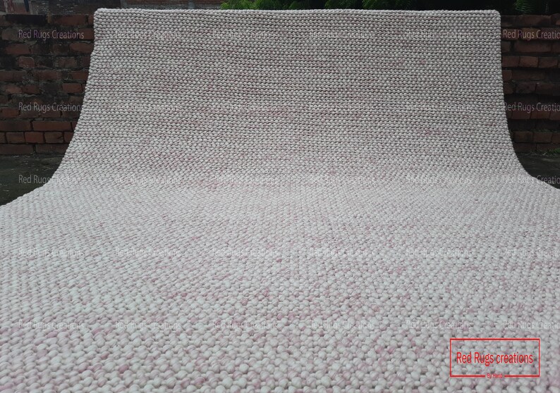 May include: A close-up of a white and pink textured rug. The rug is made of small, round balls of yarn. The rug is folded in half and the bottom edge is visible. The text "Red Rugs creations" is visible in the lower right corner of the image.