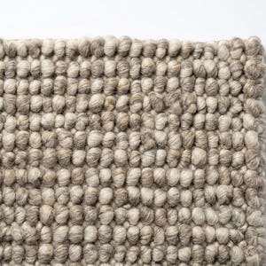 May include: A rectangular rug featuring a dense, hand-knotted texture composed of thick, rounded yarn loops. The surface displays a variegated color palette of neutral beige, soft taupe, and creamy off-white tones arranged in a uniform pattern.