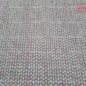 May include: Close-up of a white and pink woven rug with a textured surface. The rug is made of small, round balls of yarn that are woven together to create a unique and interesting pattern. The rug is perfect for adding a touch of texture and colour to any room.