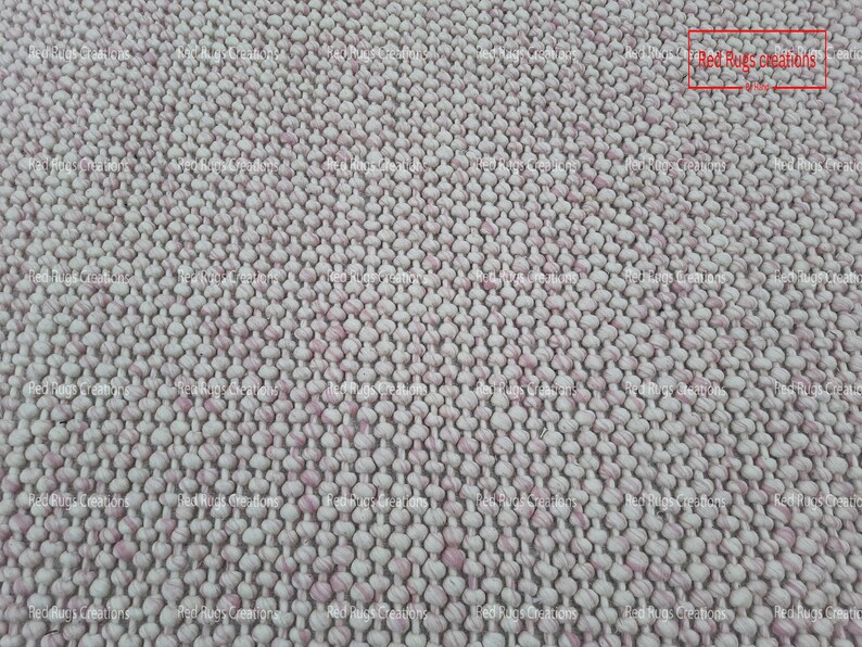 May include: Close-up of a white and pink woven rug with a textured surface. The rug is made of small, round balls of yarn that are woven together to create a unique and interesting pattern. The rug is perfect for adding a touch of texture and colour to any room.