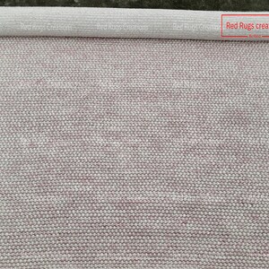 May include: A rolled up white and pink rug with a woven texture. The rug is labelled "Red Rugs creations".