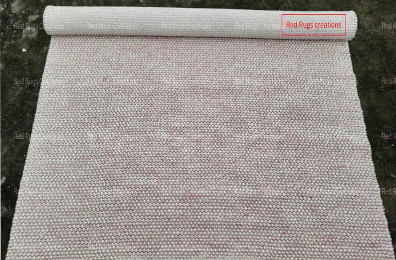 May include: A rolled up white and pink rug with a woven texture. The rug is labelled "Red Rugs creations".