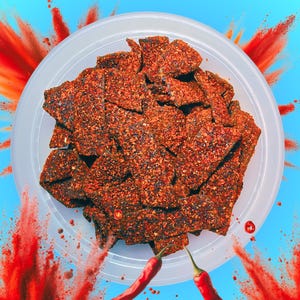 May include: A white plastic container filled with spicy, dark brown, crunchy, chili-coated snacks. The container is surrounded by red chili powder and red chili peppers.