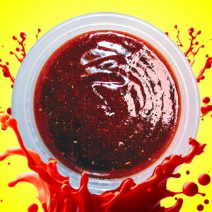 May include: A clear plastic container filled with a thick, red sauce. The sauce has a glossy finish and is surrounded by red splatters on a yellow background.