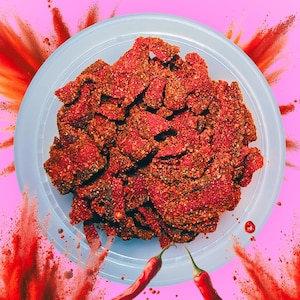 May include: A white bowl filled with red, spicy, dried fruit pieces. The bowl is on a pink background with red powder splattered around it. Two red chili peppers are visible in the background.