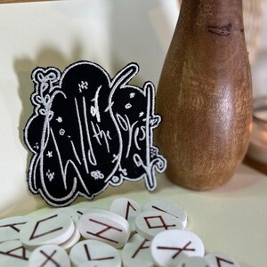 Pode incluir: Um remendo bordado em preto e branco com o texto "Wild of the Forest" rodeado de estrelas e outros desenhos inspirados na natureza. O remendo está sobre uma superfície branca com uma coleção de pedras brancas com runas vermelhas.