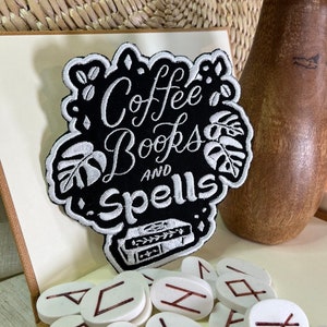 以下が含まれることがあります： 白い文字で「Coffee Books and Spells」と書かれた黒い刺繍パッチ。テキストの下には小さな刺繍された本があります。パッチは、赤いルーンが書かれた白い石のある白い表面にあります。
