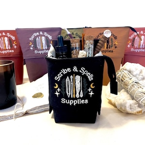 Może przedstawiać: Czarna płócienna torba z napisem "Scribe & Spells Supplies", wypełniona różnymi przedmiotami. Za nią znajduje się kilka innych toreb w odcieniach czerwieni, fioletu i brązu. Obecna jest również zapalona świeca i szałwia.