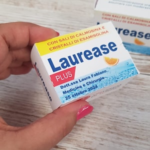 Può includere: Una scatola bianca del farmaco "Laurease PLUS" è tenuta in mano. La scatola ha accenti blu e gialli, con testo in italiano. La scatola include il nome di un medico e una data.