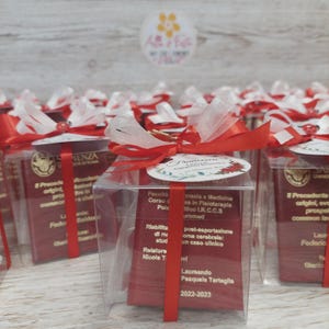 Può includere: Scatole di plastica trasparente legate con nastro rosso e tulle bianco, ognuna contenente una carta rossa con testo. Le scatole sono decorate con un piccolo tag rotondo e un fiocco rosso. Le scatole sono probabilmente bomboniere o regali.