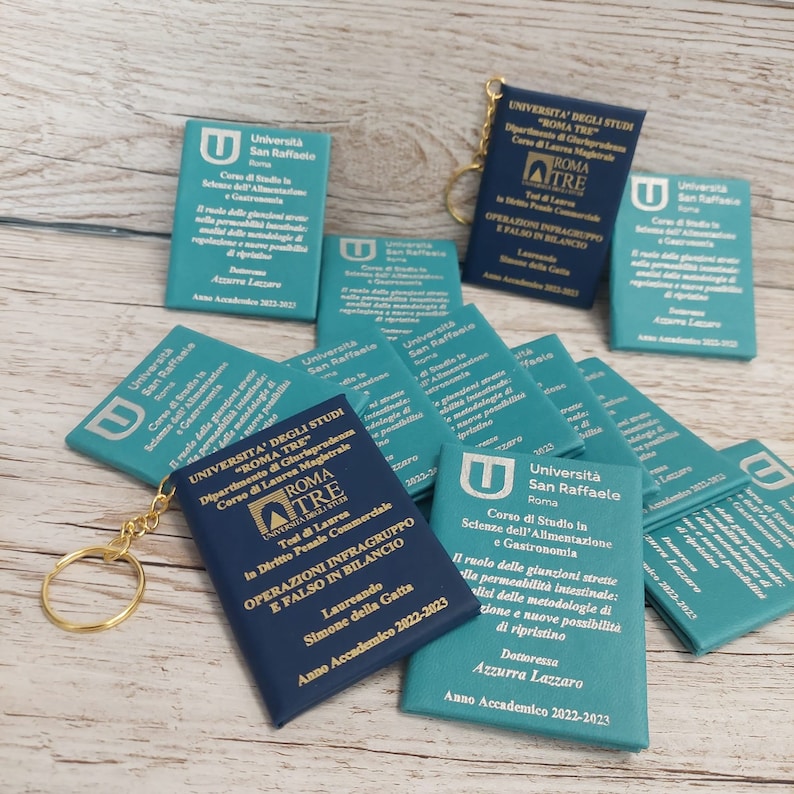 May include: A close-up of a blue and teal keychain with a small book attached. The book has the text "Universita' degli Studi "Roma Tre" Dipartimento di Giurisprudenza Corso di Laurea Magistrale" and "ROMA TRE" in gold lettering. The book also has the text "OPERAZIONI INFRAGRUPPO E FALSO IN BILANCIO" in gold lettering. The keychain is attached to a gold key ring.