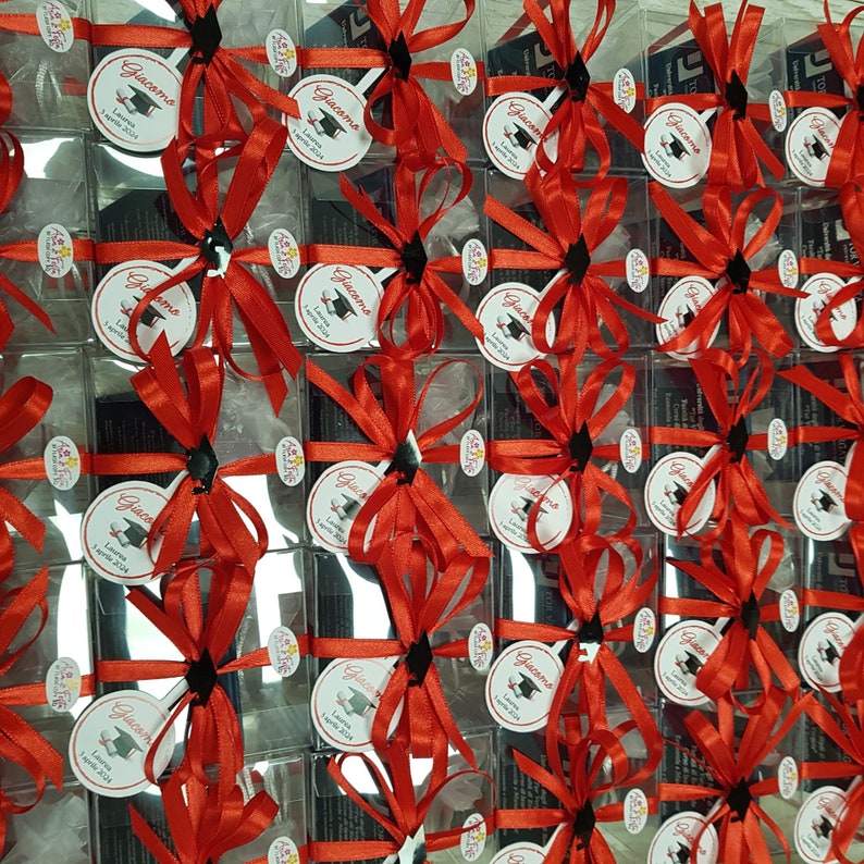 May include: A collection of small, clear boxes tied with red satin ribbon. Each box contains a small, silver mirror with a white label that reads "Giacomo Laurea 2024".