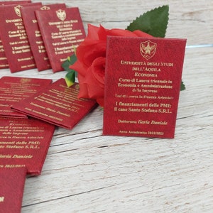 May include: A red booklet with gold lettering that reads "Universit&agrave; degli Studi dell'Aquila Economia" and "Corso di Laurea triennale in Economia e Amministrazione delle Imprese". The booklet also includes the text "I finanziamenti delle PMI: il caso Santo Stefano S.R.L." and "Dottoressa Ilaria Daniele" with the academic year "2022/2023".