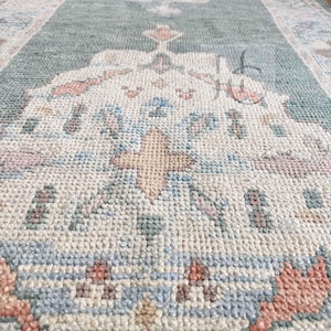 May include: A rectangular, patterned rug with a central design in shades of green, cream, and peach. The rug features a repeating floral motif and a border with a similar color palette. The rug is made of a woven material.