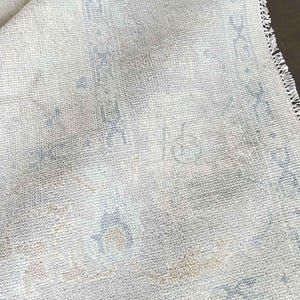 Op de afbeelding: Close-up van een opgerolde, gebroken witte vloerkleed met een vervaagd, patroonontwerp in tinten blauw en beige. Het vloerkleed heeft een getextureerd oppervlak en een gefranjerde rand, wat wijst op een vintage of traditionele stijl.