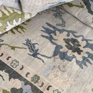 Pode incluir: Um tapete enrolado com um padrão floral detalhado. O tapete apresenta um fundo bege neutro com detalhes em azul, verde e cinza. O design inclui elementos florais e geométricos estilizados, criando uma superfície decorativa texturizada.