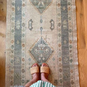 Pode incluir: Um tapete retangular de estilo vintage com um padrão central de diamante em tons de verde-azulado e bege. O tapete apresenta uma borda com motivos florais e é complementado por sandálias tecidas. O tapete está colocado sobre um chão de madeira.