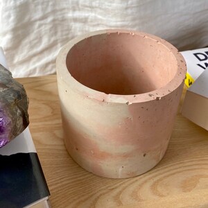 May include: A light pink and white concrete planter with a rough, textured surface. The planter is cylindrical and has a wide opening at the top.