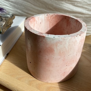 May include: A cylindrical, pink concrete planter with a rough texture. The planter has a thick rim and a smooth interior. The colour is a soft, muted pink with white accents. The planter is on a light-coloured wooden surface.