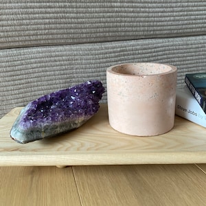 May include: A wooden tray displays a large amethyst geode and a small, cylindrical, peach-coloured concrete planter. A book titled "Steve Jobs" is partially visible. The items are arranged on a light wood surface, creating a minimalist aesthetic.