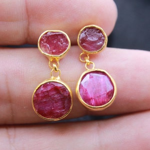 May include: Gold-toned earrings featuring two ruby gemstones per earring. The top stone is a rough-cut ruby, while the bottom is a polished, round ruby. The earrings are connected by a small gold chain.