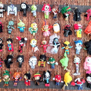 Può includere: Una collezione di portachiavi fatti a mano con vari personaggi. I portachiavi sono realizzati in filato e tessuto, con una varietà di colori e design. Alcuni portachiavi raffigurano supereroi, altri personaggi fantasy.