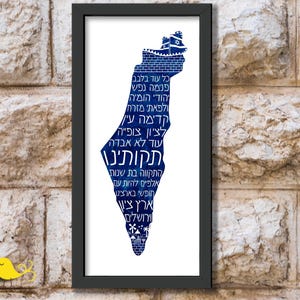 Puede incluir: Un estampado enmarcado que presenta un mapa azul de Israel, lleno de texto hebreo. El mapa está sobre un fondo blanco dentro de un marco negro. La obra de arte se exhibe en una pared de piedra texturizada.