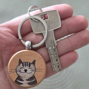 Könnte beinhalten: Ein Holz-Schlüsselanhänger mit Katze, einem silbernen Schlüsselring und einem silbernen Schlüssel. Der Schlüsselanhänger zeigt ein handgemaltes Katzen-Gesicht mit grauen Streifen und einem lächelnden Ausdruck. Der Holz-Kreis ist hellbraun.