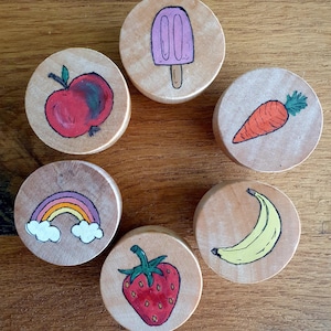 Könnte beinhalten: Sechs Holz Kreise mit verschiedenen bemalten Bildern darauf. Die Bilder sind ein roter Apfel, ein lila Eis am Stiel, ein Regenbogen, eine Banane, eine Erdbeere und eine Karotte.