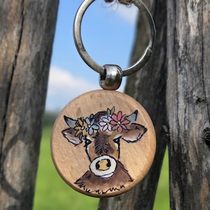 Könnte beinhalten: Ein Holzschlüsselanhänger mit handgemalter Kuh mit Blumenkranz. Die Kuh ist braun und weiß, mit schwarzer Umrandung. Oben ist ein silberner Metallring befestigt.