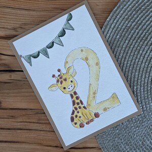 Könnte beinhalten: Eine rechteckige Grußkarte mit weißem Hintergrund und einer Aquarellillustration einer gelben Giraffe mit braunen Flecken, die die Zahl zwei bildet. Über der Giraffe befindet sich eine Girlande aus grünen dreieckigen Wimpeln.