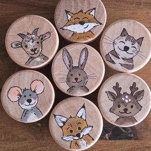 Könnte beinhalten: Sieben Holzscheiben, jede handbemalt mit einem anderen Tier. Die Tiere umfassen eine Ziege, einen Fuchs, eine Katze, ein Kaninchen, eine Maus, ein Reh und einen weiteren Fuchs. Die Illustrationen sind im Cartoon-Stil gehalten.