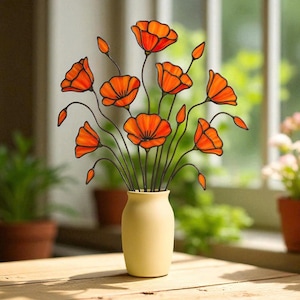 May include: A decorative arrangement of orange poppy flowers in a cream-colored vase. The flowers have black stems and are set against a blurred background. The vase sits on a wooden table, with potted plants visible.