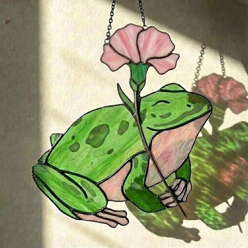 May include: Stained glass frog holding a pink flower. The frog is green with darker green spots and pink accents. The flower is pink with a green stem. The piece is designed to hang, with a chain at the top.