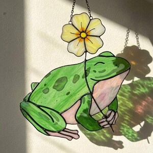 May include: Stained glass frog suncatcher with a yellow flower. The frog is green with darker green spots and a pink belly. The flower is yellow with white accents. The suncatcher hangs from a chain.