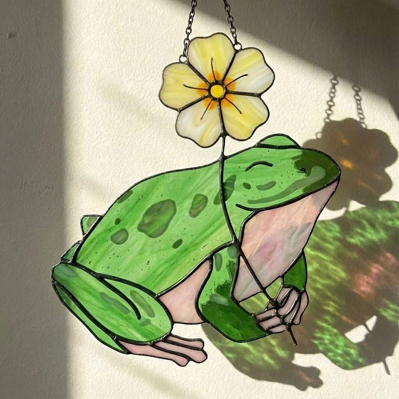 May include: Stained glass frog suncatcher with a yellow flower. The frog is green with darker green spots and a pink belly. The flower is yellow with white accents. The suncatcher hangs from a chain.