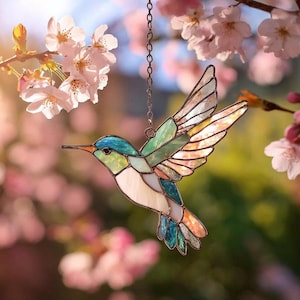 Puede incluir: Un colibrí de vidriera con tonos azules, verdes, blancos y naranjas cuelga de una cadena. El pájaro está representado en pleno vuelo, sobre un fondo borroso de flores rosas y sol.