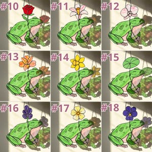 May include: A collection of stained glass frog suncatchers, each featuring a different flower. The frogs are green with white bellies and are suspended by a chain. The flowers include a red rose, white lily, pink orchid, orange lily, yellow daffodil, and more.