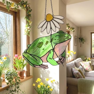 May include: A stained glass frog with a daisy, hanging from a chain. The frog is green with pink accents and a smiling face. The daisy has a yellow center and white petals. The artwork is displayed in a sunlit room with plants.