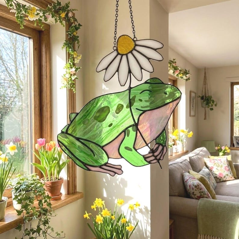 May include: A stained glass frog with a daisy, hanging from a chain. The frog is green with pink accents and a smiling face. The daisy has a yellow center and white petals. The artwork is displayed in a sunlit room with plants.