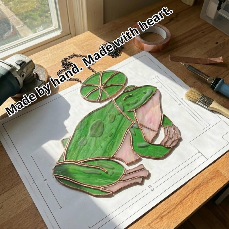 May include: A stained glass frog with a lily pad pendant. The frog is green with pink accents and copper edging. The text "Made by hand. Made with heart." is visible. The artwork is on a white paper with measurements.