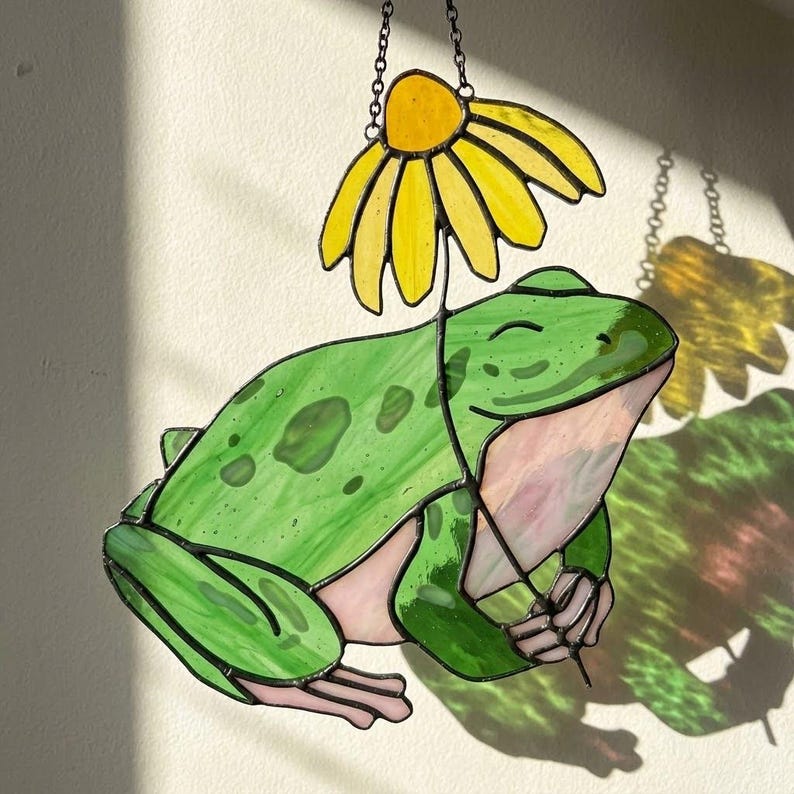 May include: A stained glass frog with a yellow daisy umbrella. The frog is green with darker green spots and pink accents. The daisy has yellow petals and a yellow center. The piece hangs from a chain.